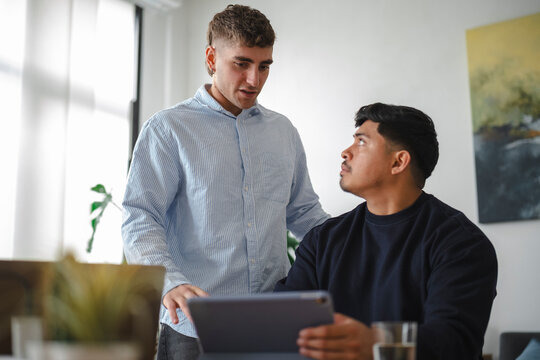 Two young men collaborate on a project in a modern living room. One man stands and talks while the other sits holding a digital tablet, listening to his colleague.