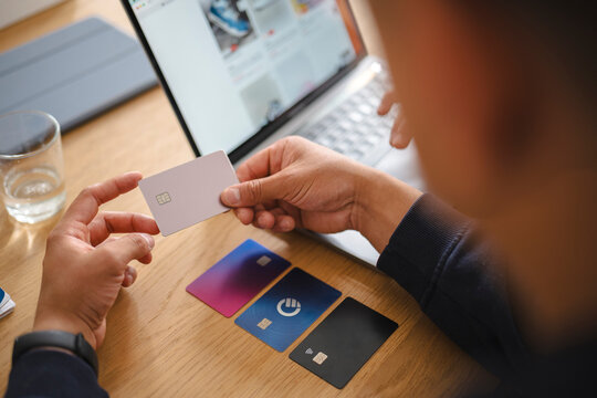 A person sitting at a wooden desk with a laptop holds a credit card in their hands while making an online purchase. Several other payment cards lie on the table.
