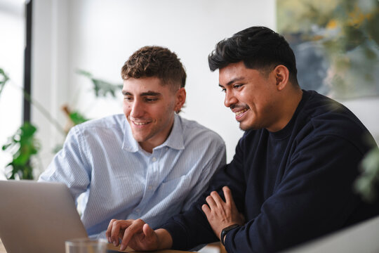 Two young men collaborate on a laptop in a bright office. They smile while reviewing a project together, showing teamwork and engagement in a modern workspace.