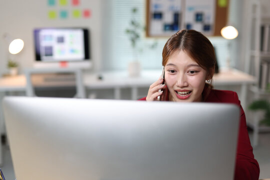 Young Asian businesswoman talking on smartphone and smiling while working at desk in a corporate office.
