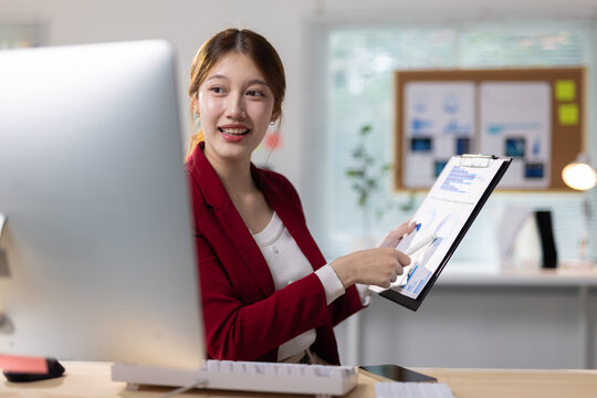 Smiling Asian business woman reviewing financial documents on a clipboard, working efficiently at her office desk.