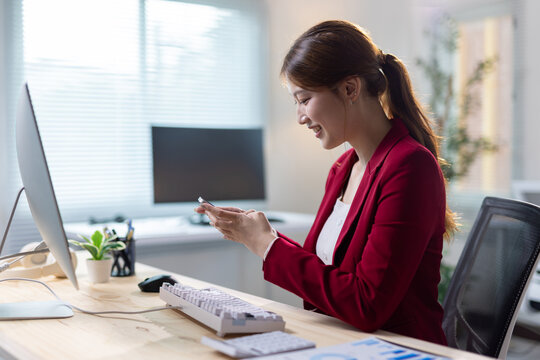 Businesswoman smiling and using smartphone at office. Asian businesswoman smiling, using a smartphone while working at a modern office desk, digital communication and productivity.
