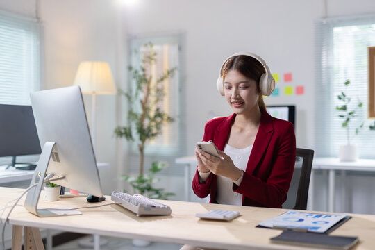 Asian business woman multitasking using smartphone and headphones. Young Asian business woman working in office, wearing headphones and using smartphone while sitting at desk.