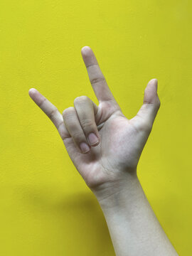 Close up of a hand making the rock and roll sign against a bright yellow background, representing music culture, freedom, youth expression, and alternative lifestyle.