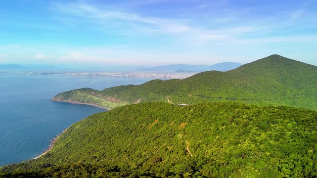 Sunlit green hills meet the blue sea on Son Tra Peninsula near Da Nang, Vietnam, with distant coastline and scattered buildings beneath a clear daytime sky