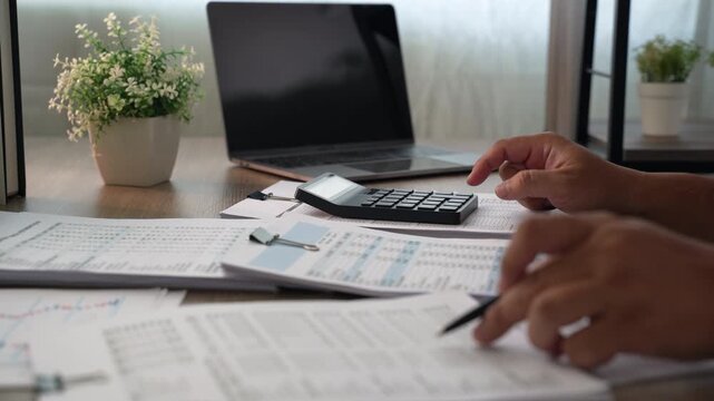Office professional reviewing financial documents with calculator, laptop and pen, comparing report figures, expenses and budget data for business accounting analysis and planning.