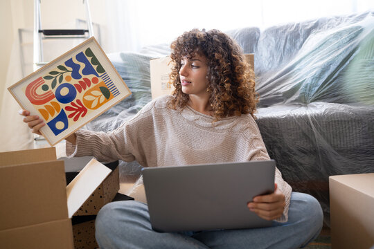 White woman holding artwork and laptop surrounded by moving boxes