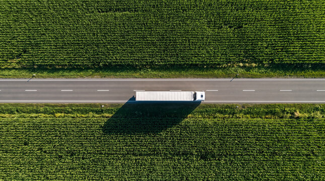 White semi-truck driving on a scenic highway surrounded by lush green fields from an aerial view