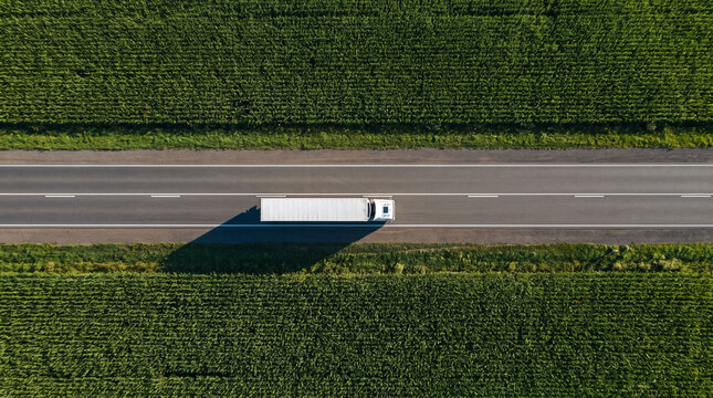Aerial top down view of white semi truck driving on asphalt road through green agricultural fields