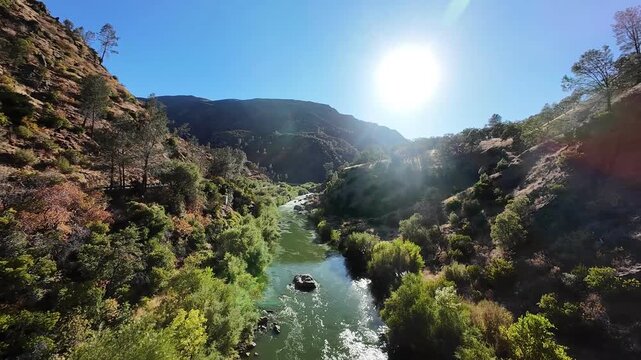 First-person view drone flying at high speed through a sunny canyon, following the beautiful Cache Creek river surrounded by lush green trees and arid hills under a clear blue sky in California