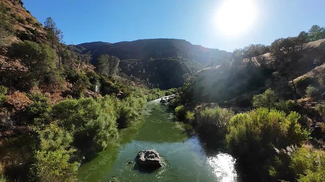 Cinematic aerial footage shows lush greenery and glistening water of Cache Creek winding between sunlit hills, with dramatic shadows and natural textures under clear blue sky, captured by FPV drone.