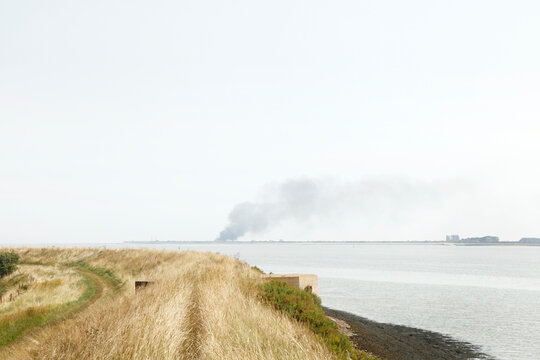 Coastal Estuary Landscape With Distant Smoke Plume Rising Over Far Shoreline