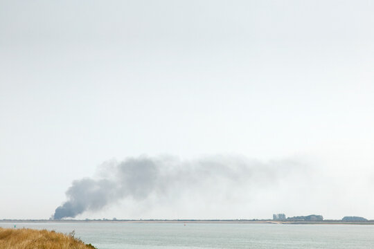 Dark Smoke Plume Rising From Coastal Fire Across Calm Water Under Overcast Sky