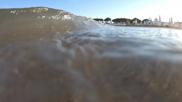 Half-underwater view of a small wave breaking over the sandy shore, with closed parasols and pine trees on the beach, French Riviera, France.