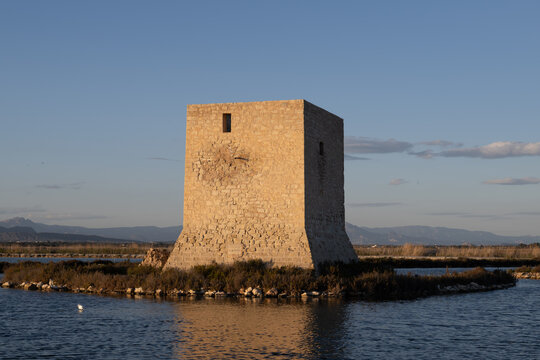 Quadratischer Steinturm auf kleiner Schilfinsel im ruhigen Wasser; warmes Seitenlicht betont die gemauerte Fassade. Flacher Horizont mit Bergen, klarer Himmel, Reflexion im Wasser.