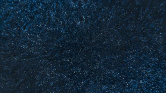 Blue particles flow through rippling curved formations across dark navy background, creating shifting waves of luminous specks and layered abstract patterns with subtle depth