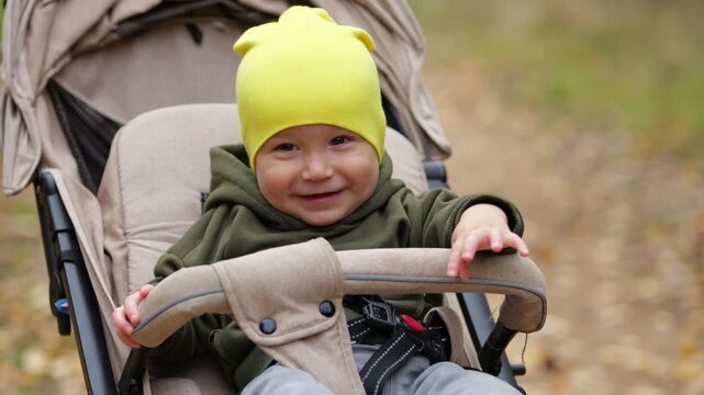 Cute Infant Rides Warm Buggy During Autumn Park Excursion. Joyful Baby Girl Wearing Bright Beanie Smiles Broadly While Seated Toddler Enjoys Gentle Forest Stroll Among Falling Gold Foliage.