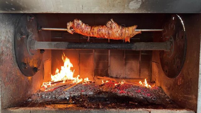 Whole suckling pig roasting on a rotating spit over hot charcoal flames in a traditional outdoor oven. Crispy whole roasted piglet preparing traditional barbecue feast