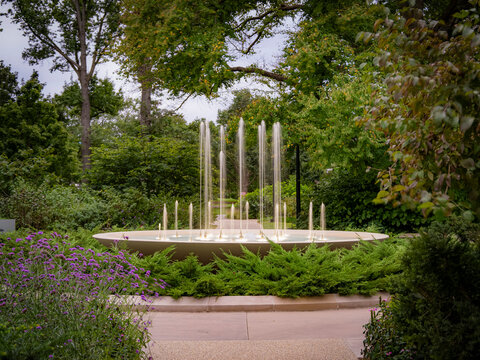 Water fountain spraying jets in an outdoor garden park with green trees purple flowers and stone paving