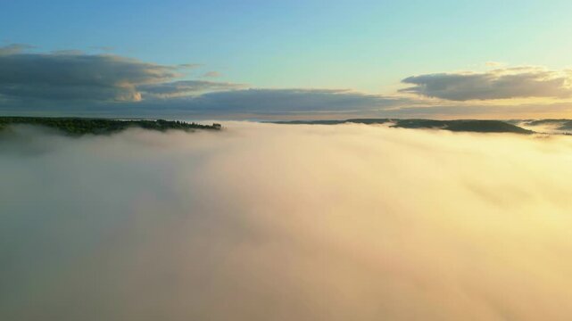 High-altitude aerial shot showing a thick blanket of fog rolling over forest hills, illuminated by warm sunlight at sunrise under a blue sky.