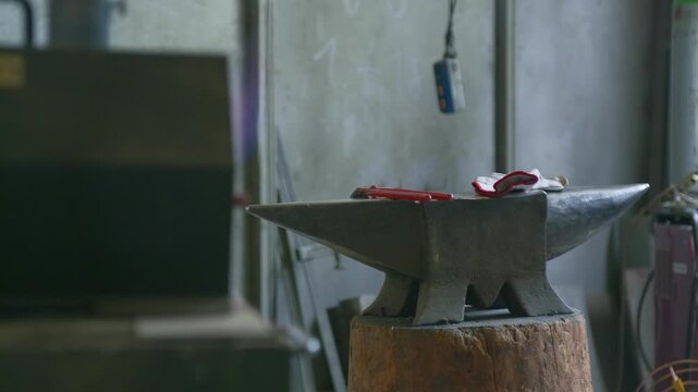 Anvil blacksmith workshop tools forge resting on a wooden stump with pliers and gloves in a traditional smithy. Static tripod shot of empty industrial metalworking workspace.