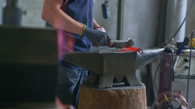 Blacksmith cleaning hot glowing metal with a wire brush on an anvil inside a forge workshop. Detail of craftsman hands working with glowing iron, static tripod shot.