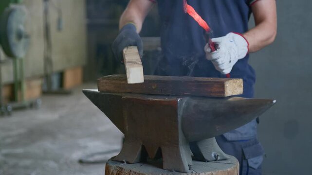 Blacksmith hammering hot glowing metal on a wooden block placed on an anvil inside a forge workshop. Detail of craftsman hands forging glowing iron with a hammer, static tripod shot.