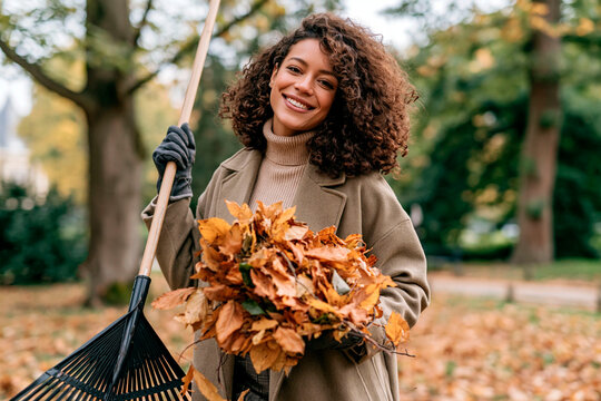 Young woman gathering fallen autumn leaves with rake in park, smiling gardener doing seasonal outdoor cleanup and yard maintenance