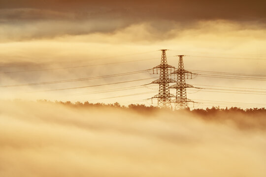 High voltage power transmission towers in thick mist forest field under golden morning sunlight. Industrial electric grid wire landscape