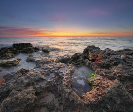 Beautiful rocky sea coast under vibrant sunset sky with long exposure smooth water effect. Twilight ocean shore landscape