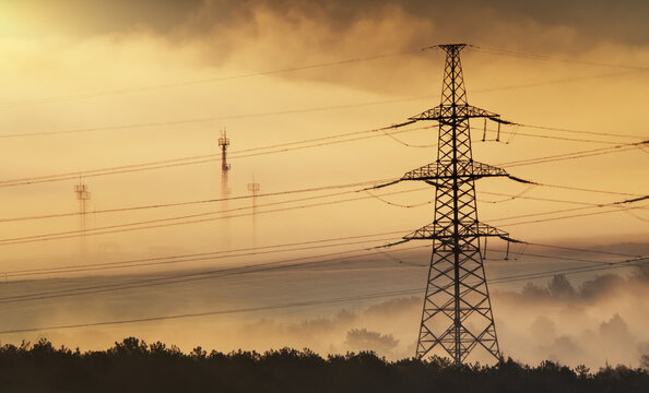 High voltage power transmission pylon with telecommunication cellular towers in thick fog under golden sunrise sky. Industrial electric grid landscape