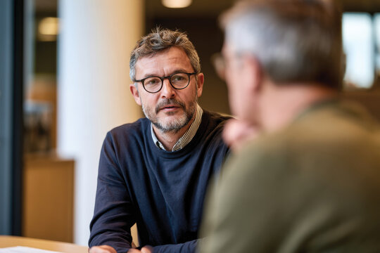 A man in a navy sweater attentively engages in a conversation with a colleague, embodying professionalism and collaboration in a contemporary office environment.