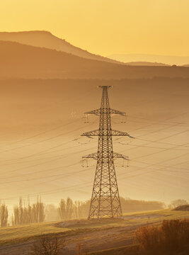 High voltage electric power transmission pylon on grassy hill near mountain layers during golden sunrise light. Industrial grid steel tower