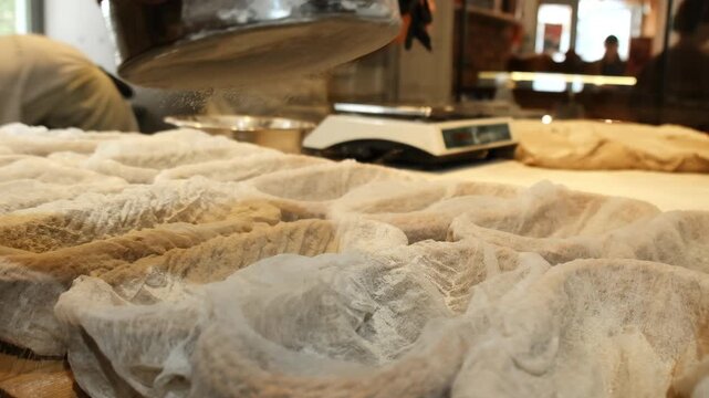 Baker gently dusts pale dough portions inside rattan baskets with fine wheat flour. Man uses small sieve to create light even coating. Slow motion
