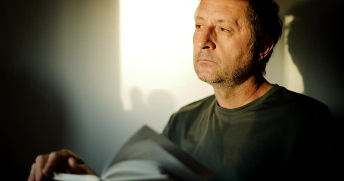 Thoughtful man holding an open book, gazing into the distance in a shaft of light with deep contrast and shadow. 4K.