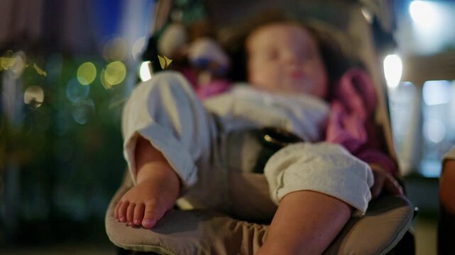 Close-up of the bare feet and legs of a baby girl relaxing in a stroller at night, her face softly blurred behind, warm bokeh lights, French Riviera, France.