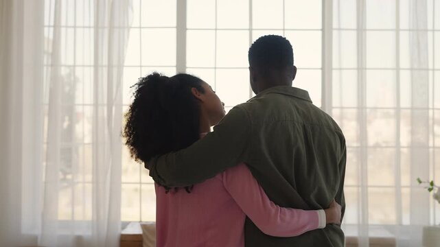Young African American couple embrace while looking out window, true love