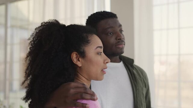 Upset young Black woman leaning on husband's shoulder, looking out window
