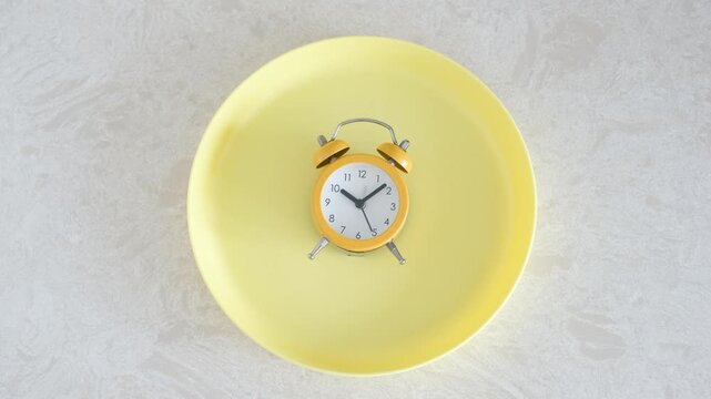 Yellow empty plate with alarm clock on gray background, intermittent fasting concept.
