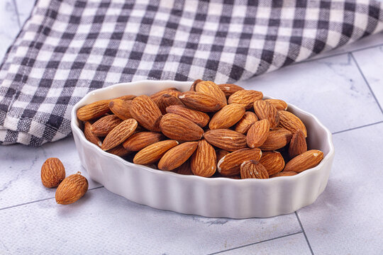 White ceramic dish filled with whole raw almonds on light gray tiled surface beside black and white checkered cloth snack food