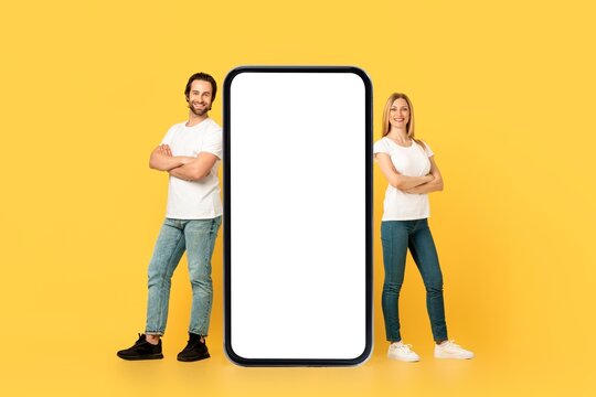 Smiling Caucasian Man And Woman Presenting Smartphone With Empty Screen, Standing Together In Studio Against Yellow Background, Promoting New App Or Product Concept, Creative Advertisement.