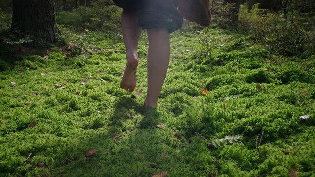 Caucasian young woman walking barefoot on green moss in woodland while carrying hiking boots in rear tracking shot. Ideal for ecotourism and grounding wellness campaigns