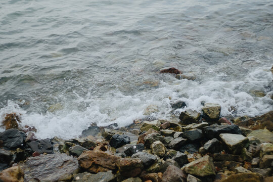 A top-down high-angle shot capturing ocean waves and white sea foam gently crashing against a rocky shore strewn with natural pebbles and stones, set in clear shallow coastal water.