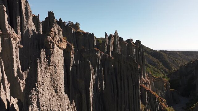 A drone glides close beside jagged earth spires at Putangirua Pinnacles. Rough grey surfaces catch the sunlight while a wooded valley recedes beyond the sharply eroded ridge.