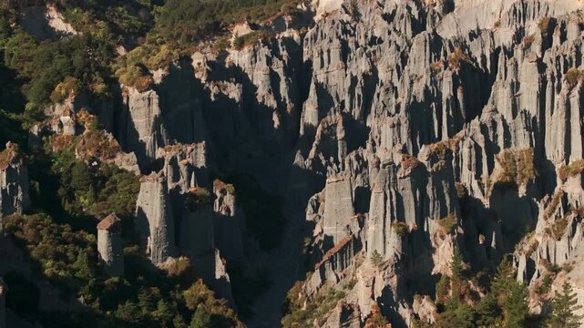 An aerial view traces the sculpted cliff face of Putangirua Pinnacles, showing rows of grey towers, scattered shrubs and green trees around a deep shaded gully in Wairarapa.