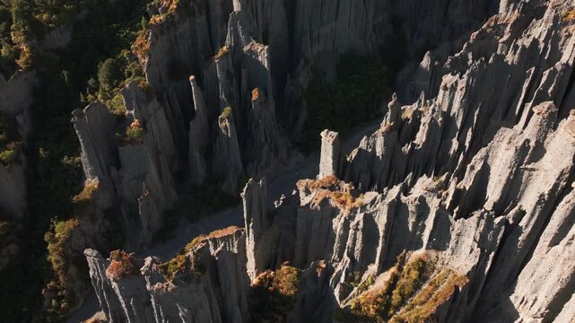An oblique drone pass follows grey earth pillars along a stony ravine at Putangirua Pinnacles. Sunlit edges, shadowed walls and small shrubs reveal the intricate eroded landscape.