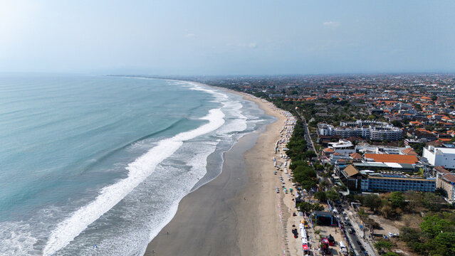 Aerial view of Kuta Beach with waves rolling onto the sandy shore, lined with hotels and resorts along the coastline in Kuta, Bali, Indonesia.