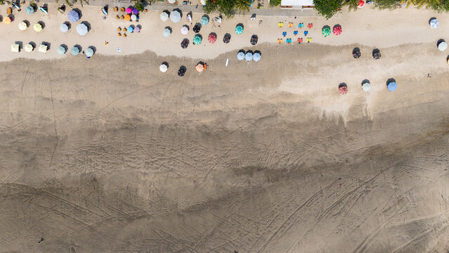 Aerial view of Kuta Beach featuring rows of colorful umbrellas on the sand with visible tire tracks and coastal textures in Kuta, Bali, Indonesia.