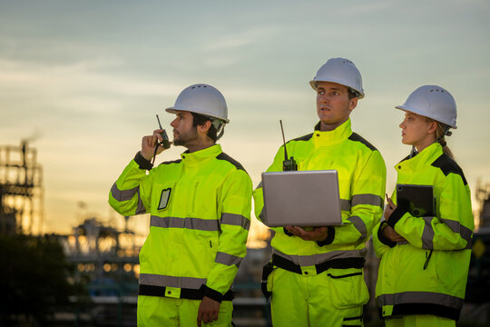 Group of industrial inspectors using laptop and radio communication at oil refinery during dusk, Industrial engineering team holding laptop, tablet and walkie talkie at refinery plant at sunset