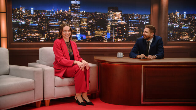 Evening Television Interview on Studio Set: Woman Guest Smiles on Armchair During Fun Talk as Male Presenter Laughs Behind Desk, Night Skyline Backdrop, Celebrity Chat Program